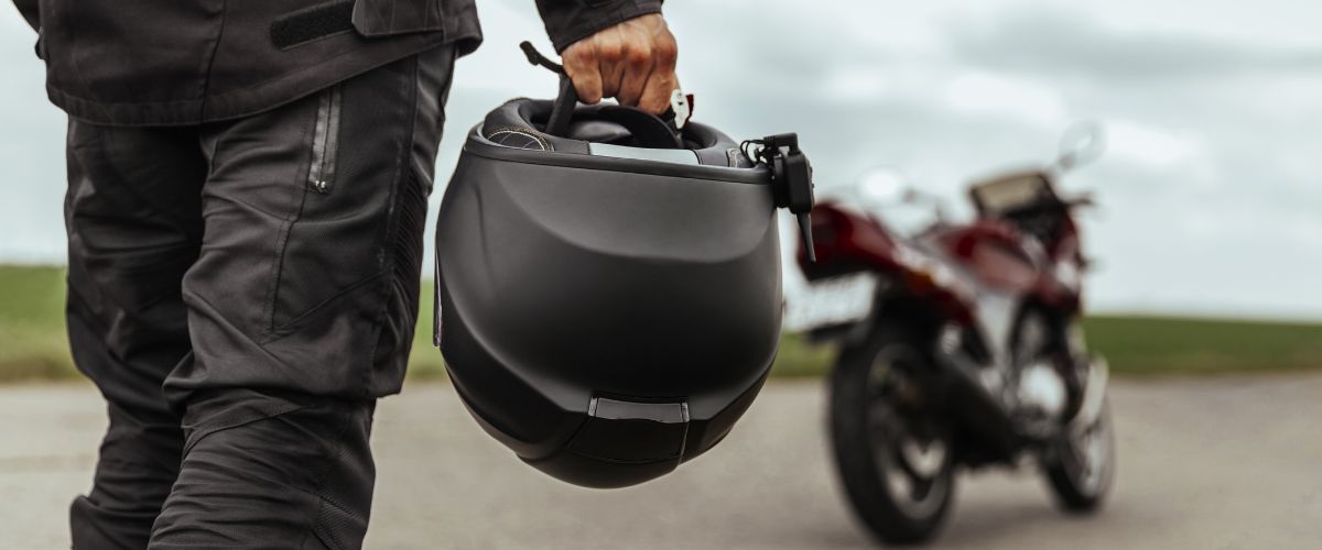 A motorcycle rider in black, holding a black helmet, walking toward his motorcycle.
