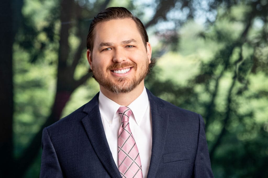 Professional headshot of smiling attorney in suit and tie posing outdoors in front of green trees.