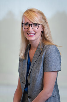 Alt text: Smiling blond woman in glasses and gray shirt, welcoming injury attorney headshot