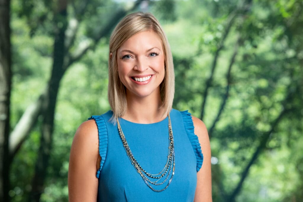 Smiling blonde woman in blue top with necklace against blurred green tree background