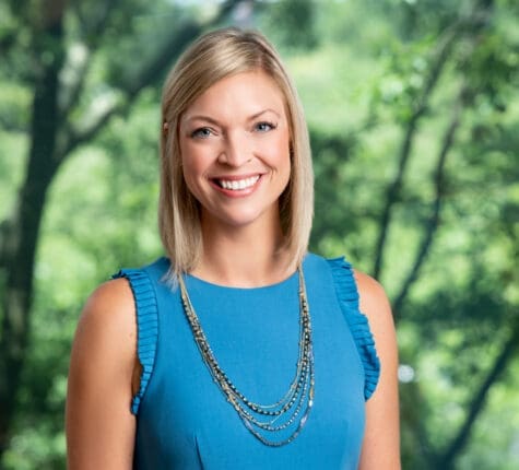Smiling blonde woman in blue top with necklace against blurred green tree background
