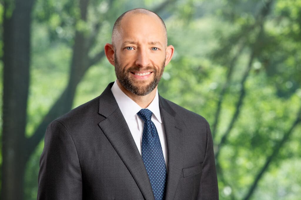 Smiling attorney headshot in front of green trees, wearing suit and blue tie, professional legal portrait
