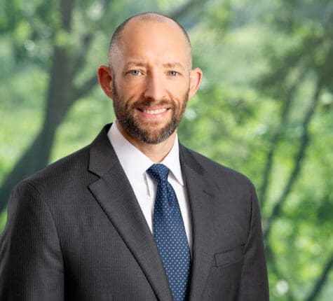 Smiling attorney headshot in front of green trees, wearing suit and blue tie, professional legal portrait