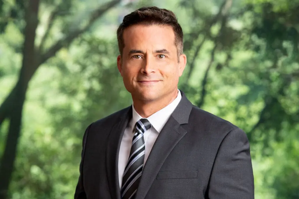 Professional headshot of a smiling man in a suit and tie in front of blurred trees, an attorney profile photo