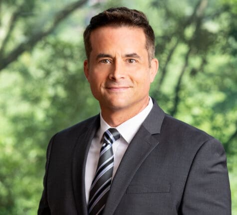 Professional headshot of a smiling man in a suit and tie in front of blurred trees, an attorney profile photo