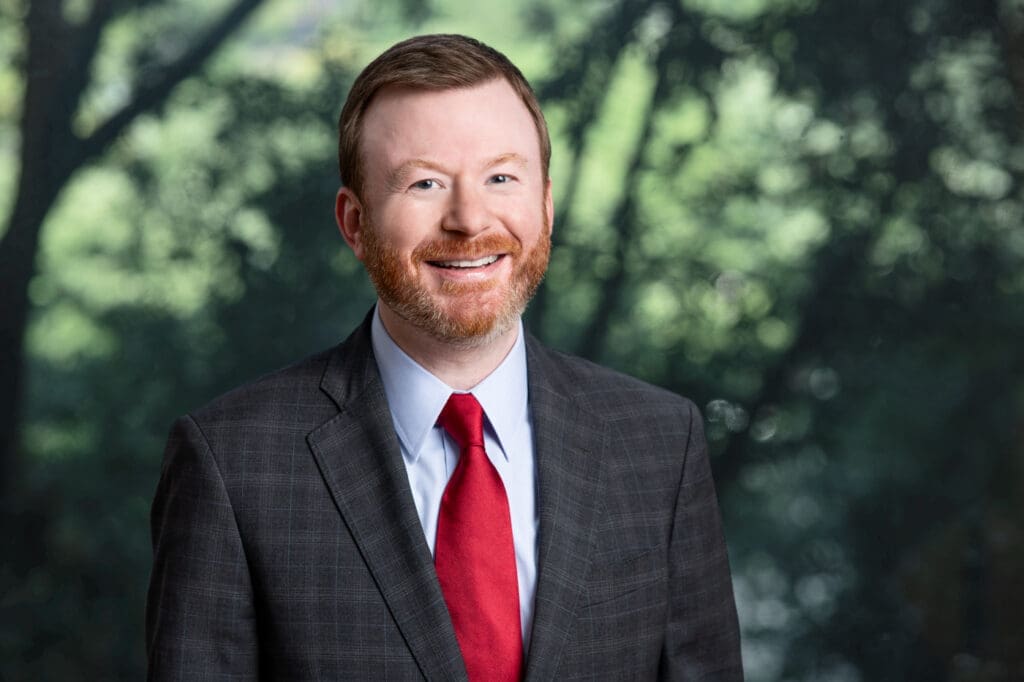 Smiling attorney with red tie and gray suit in front of blurred trees, ready to assist with legal matters.