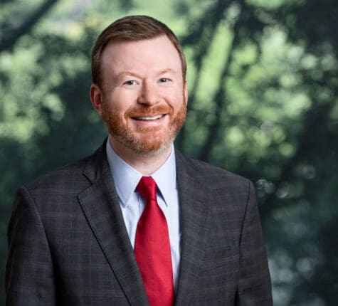 Smiling attorney with red tie and gray suit in front of blurred trees, ready to assist with legal matters.