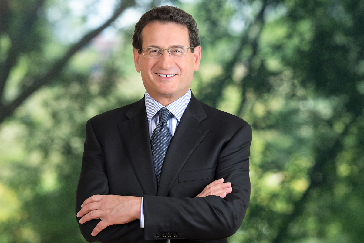 Professional headshot of smiling businessman in suit and glasses posing outdoors with blurred trees in background
