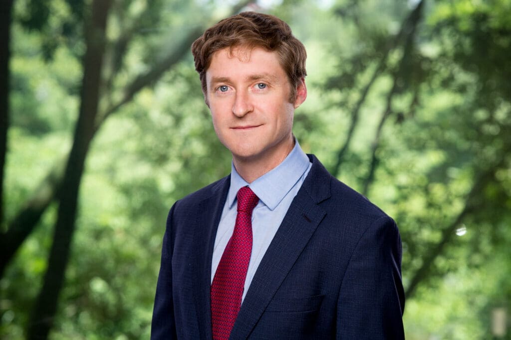Professional headshot of a smiling attorney in a blue suit and red tie surrounded by lush trees, conveying approachability and expertise for potential clients seeking legal representation.