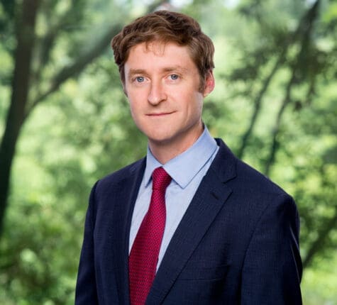 Professional headshot of a smiling attorney in a blue suit and red tie surrounded by lush trees, conveying approachability and expertise for potential clients seeking legal representation.