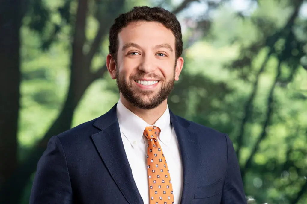 Professional headshot of smiling attorney in suit and tie with blurred trees in background