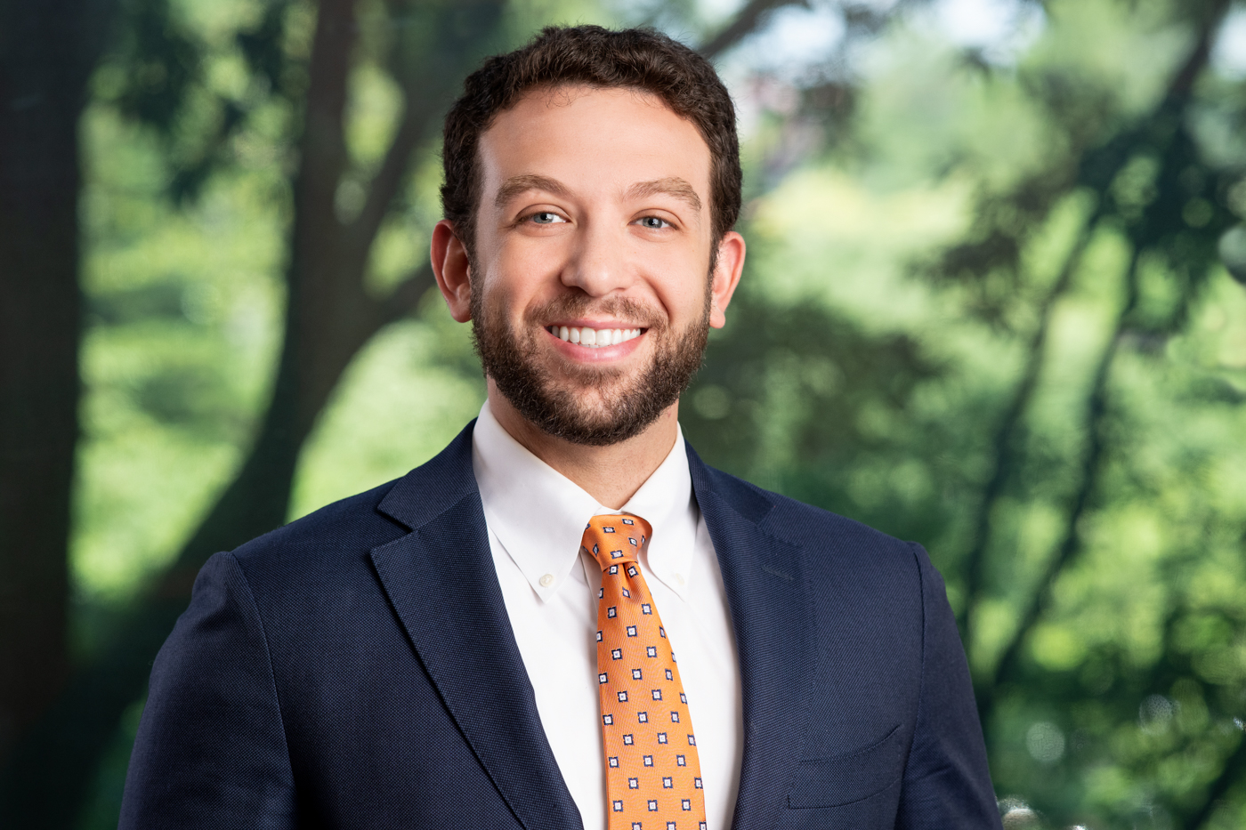 Professional headshot of smiling attorney in suit and tie with blurred trees in background