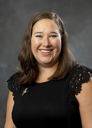 Smiling female attorney wearing black top with lace sleeves, brown hair, on grey background.