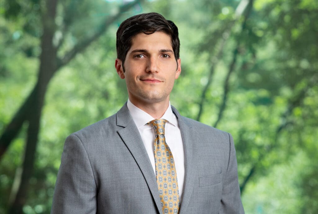 Young male professional in gray suit and patterned tie smiling outdoors in front of blurred green trees.