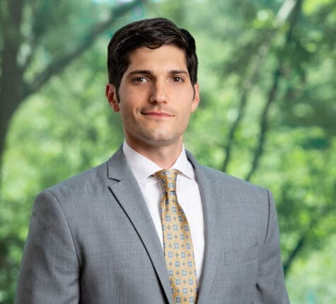 Young male professional in gray suit and patterned tie smiling outdoors in front of blurred green trees.