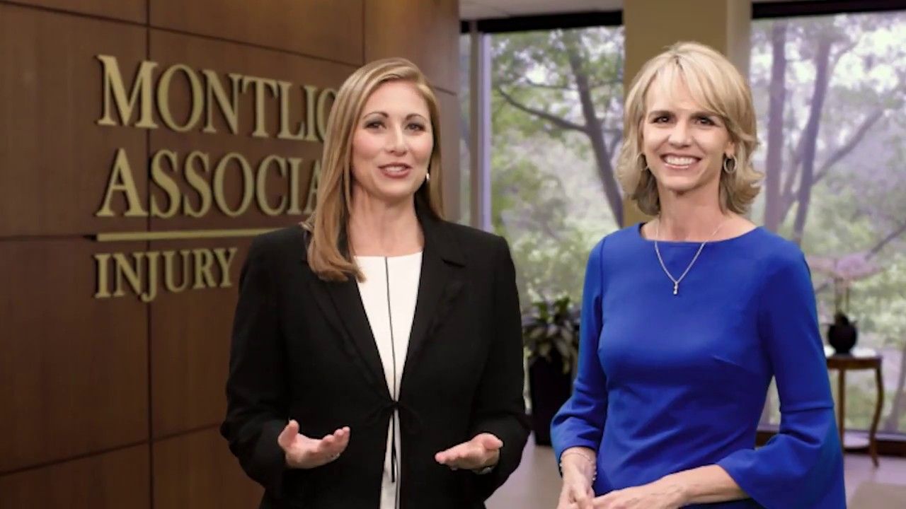 Two smiling women in business attire standing in front of a Montlick & Associates Injury law firm sign.