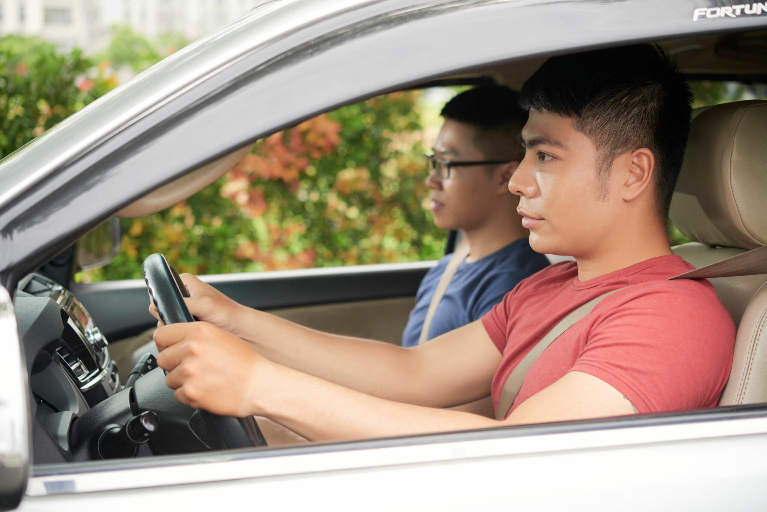 Two young men sit in a car, one driving while the other looks concerned, suggesting driver distraction.
