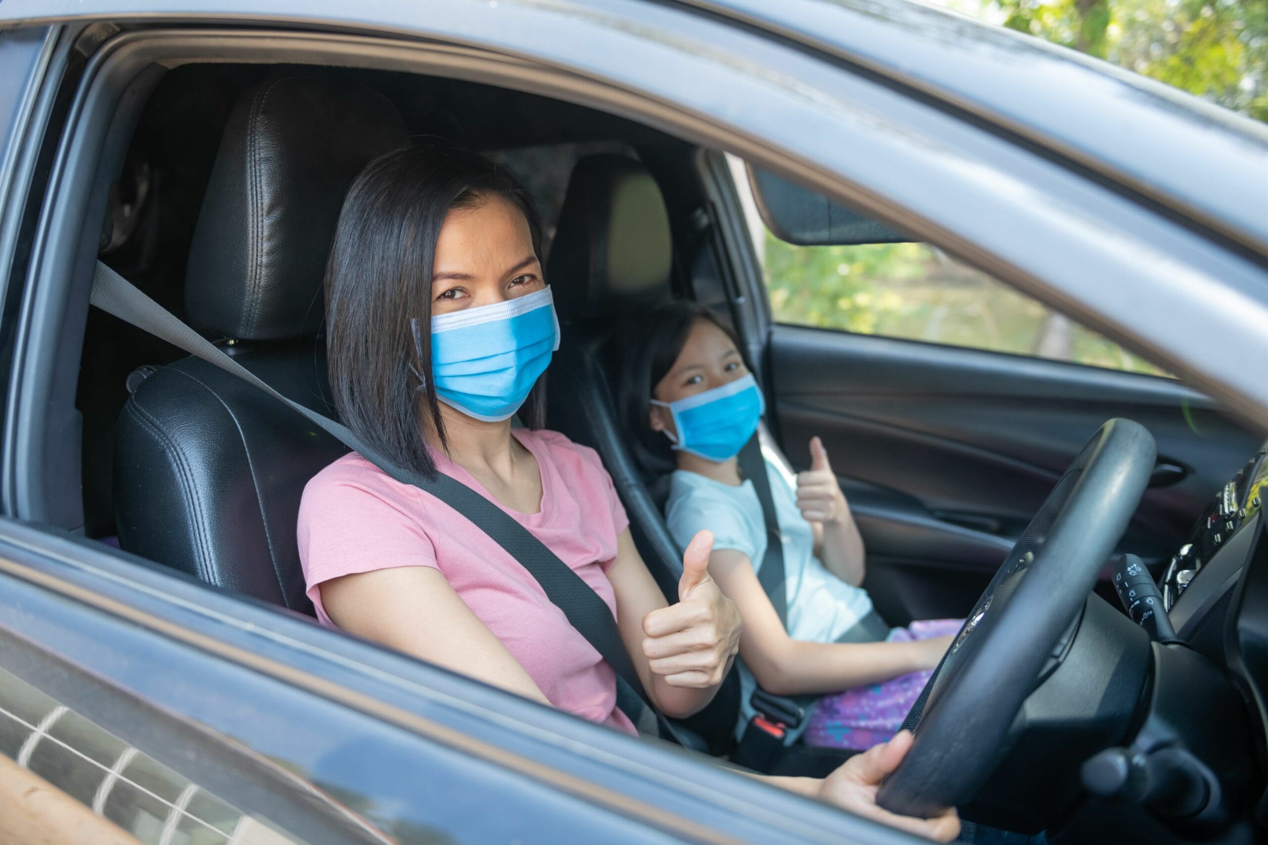 Mother and daughter wearing masks while driving in car, giving thumbs up sign. Seatbelts on for safety.