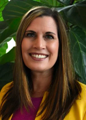 Smiling woman with brown hair in pink shirt surrounded by green plants, welcoming potential legal clients