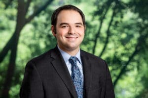Smiling attorney headshot in suit and tie against blurred green tree background