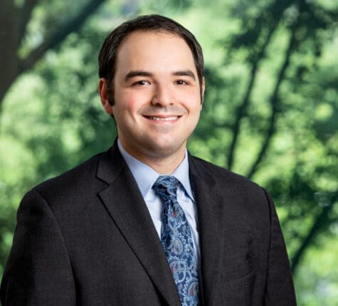 Smiling attorney headshot in suit and tie against blurred green tree background