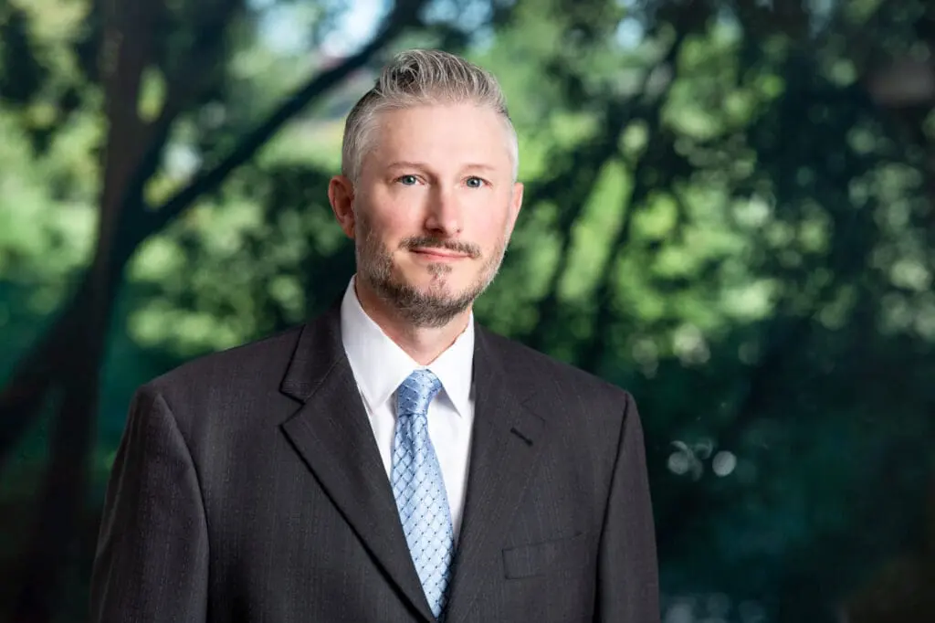 Professional headshot of a smiling businessman in a suit and tie outdoors surrounded by trees, an SEO-friendly image for an injury attorney website.