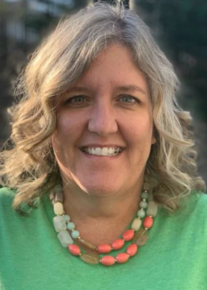 Smiling woman with blonde curly hair wearing green shirt and colorful beaded necklace, outdoors