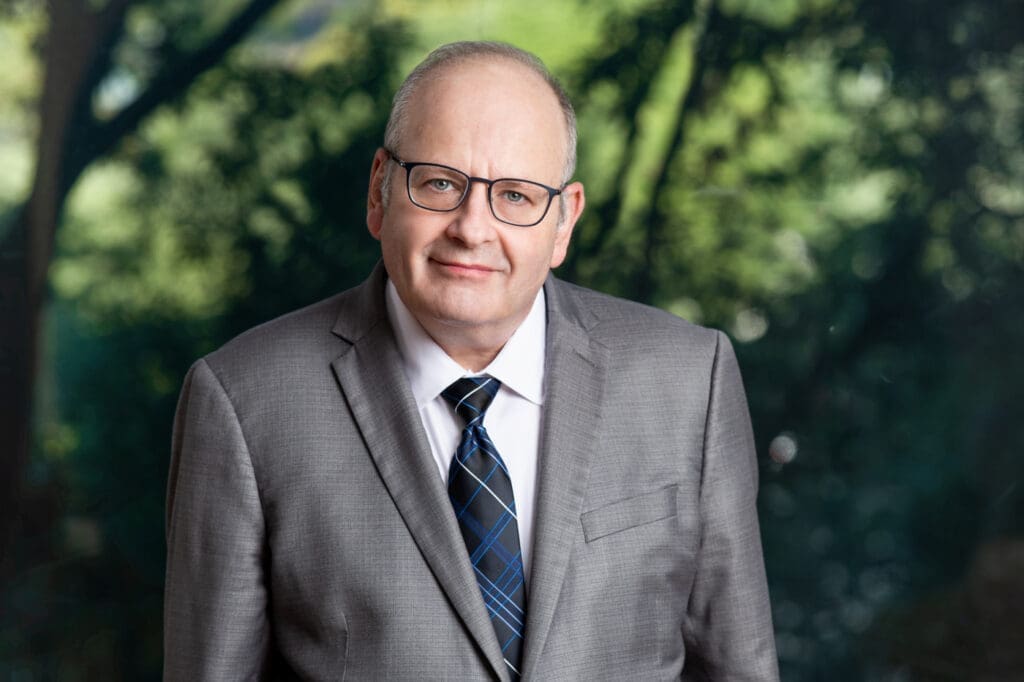 Professional headshot of a smiling senior businessman in glasses and grey suit outdoors in nature setting