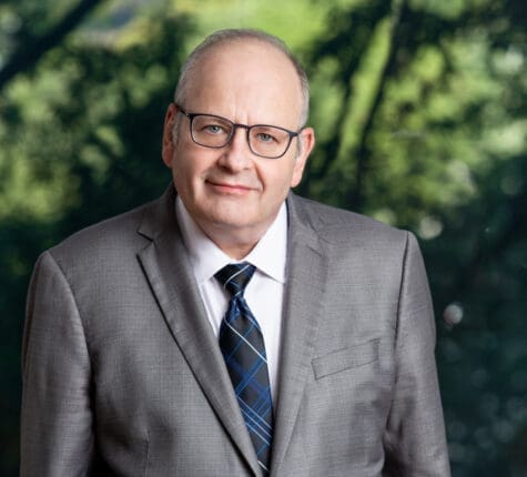 Professional headshot of a smiling senior businessman in glasses and grey suit outdoors in nature setting