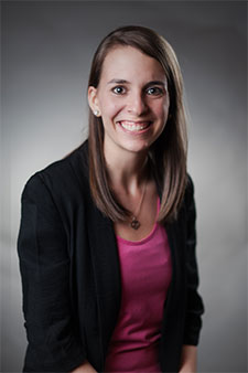 Young smiling woman with brown hair wearing maroon top and dark cardigan sweater, on gray background.