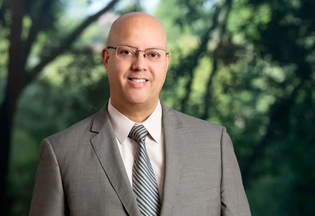 Smiling bald attorney wearing glasses and gray suit standing outdoors, ready to help with your legal needs.