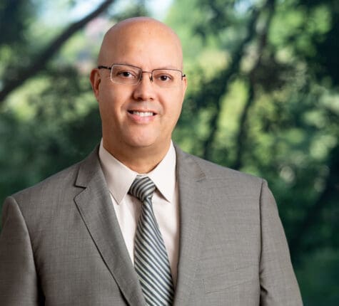 Smiling bald attorney wearing glasses and gray suit standing outdoors, ready to help with your legal needs.