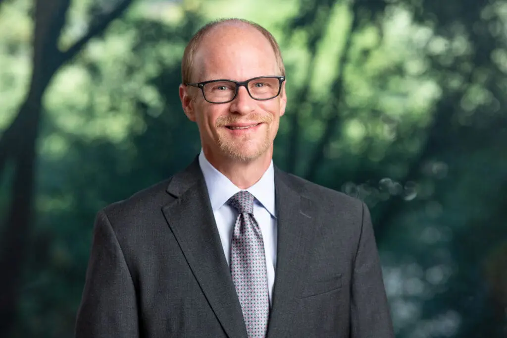 Smiling bespectacled man in suit and tie outdoors with blurred trees in background