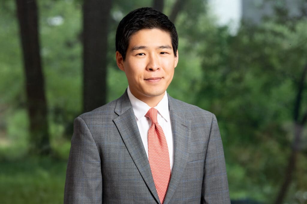 Asian businessman in grey suit and red tie smiling in forest setting, representing approachable legal expertise.