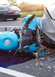 Blue motorcycle crash on road, damaged bike parts scattered on pavement, blurred car in background