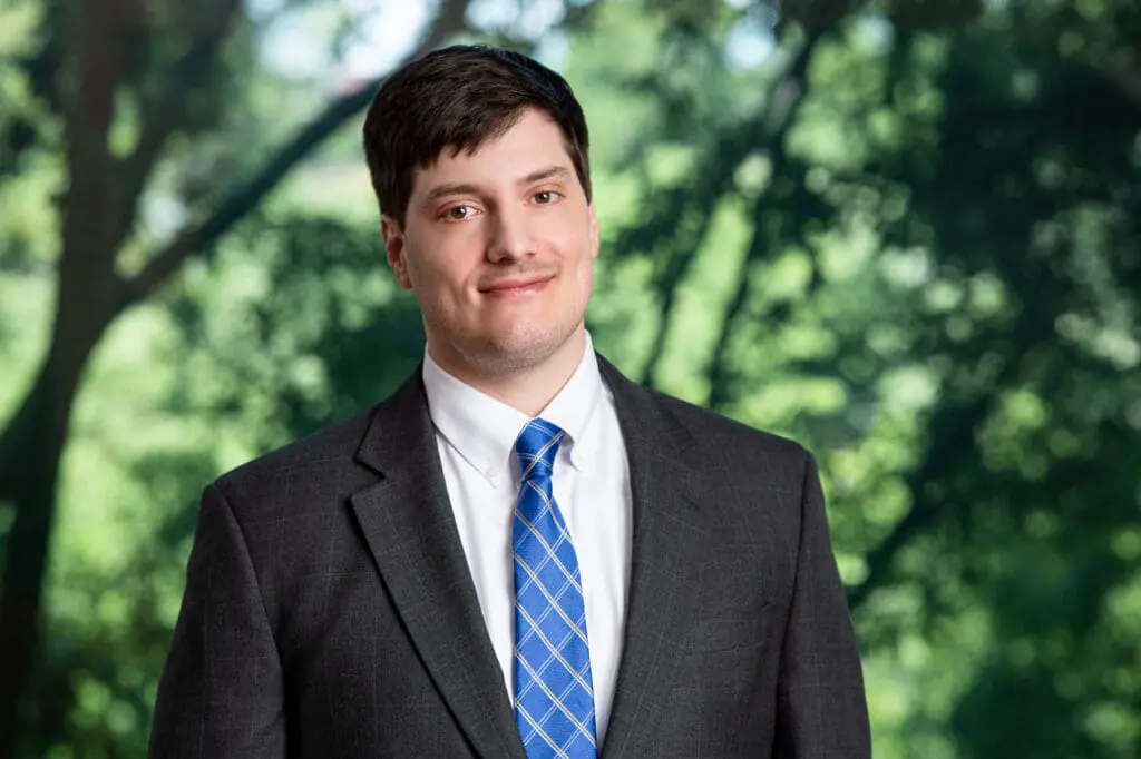 Young businessman in grey suit and blue tie smiling confidently outdoors among trees and foliage.