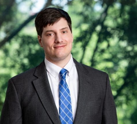 Young businessman in grey suit and blue tie smiling confidently outdoors among trees and foliage.