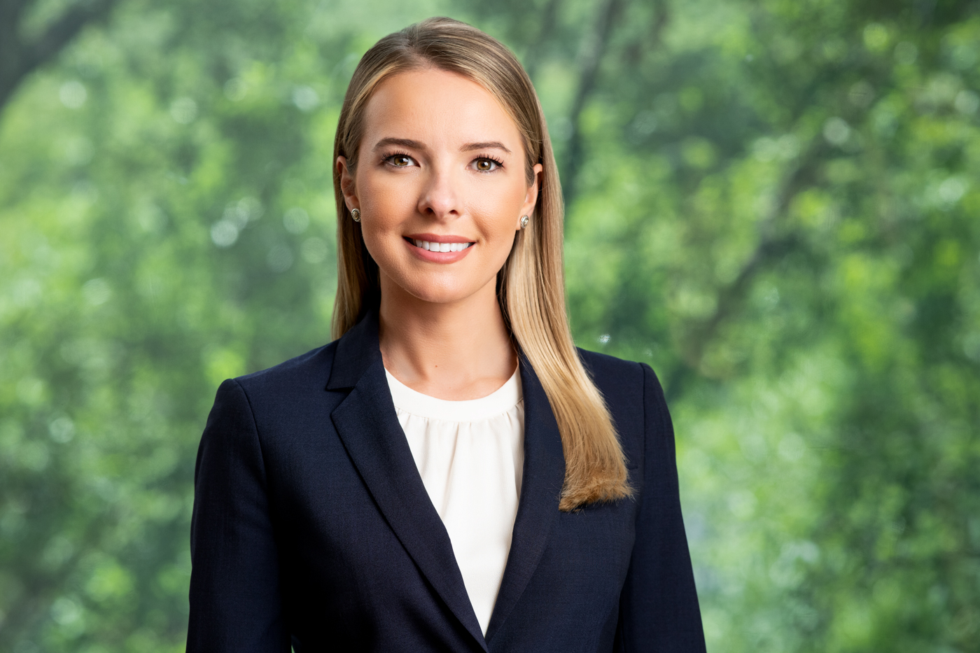 Smiling blonde female attorney in navy blazer, professional headshot with green foliage background.