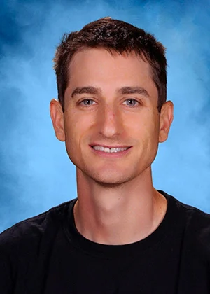 Smiling young man with short brown hair wearing a black t-shirt against a blue background portrait