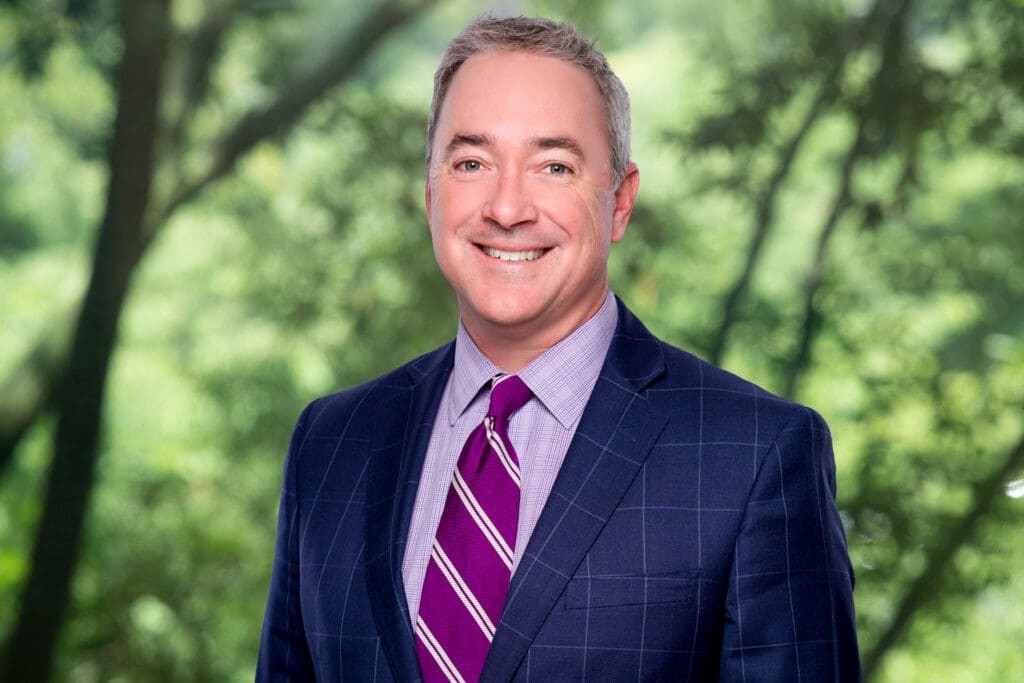 Middle-aged male attorney in blue suit and purple tie smiling warmly in front of trees