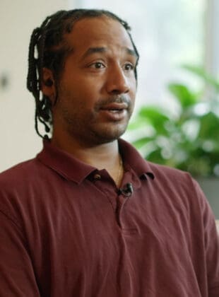 Wrong-Way Crash Injury Lawyers_ Anthony's Story Young African American man with dreadlocks gazing pensively, wearing a burgundy collared shirt, in a brightly lit room with plants in the background.