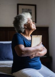 Elderly African American woman with gray hair holding picture frame, looking upward pensively