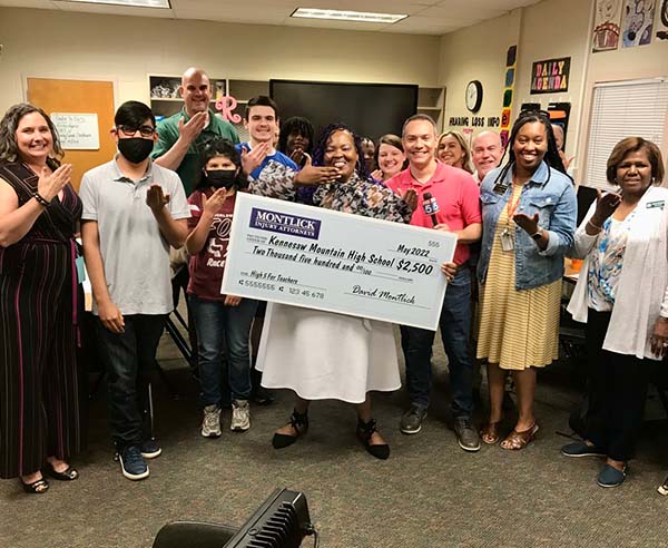 High school students and teachers hold giant check donation from personal injury law firm, smiling in classroom.