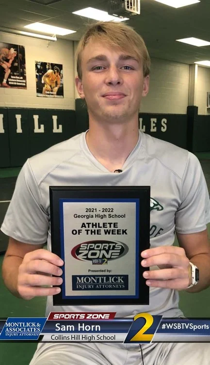 Male high school athlete holding "Athlete of the Week" award plaque in gym, smiling at camera