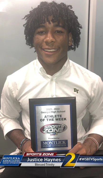 Young African American male student athlete holding an "Athlete of the Week" award smiling at the camera.
