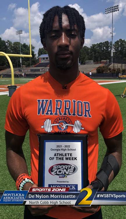 High school athlete of the week De'Nylon Morrissette standing on football field in orange Warrior jersey.