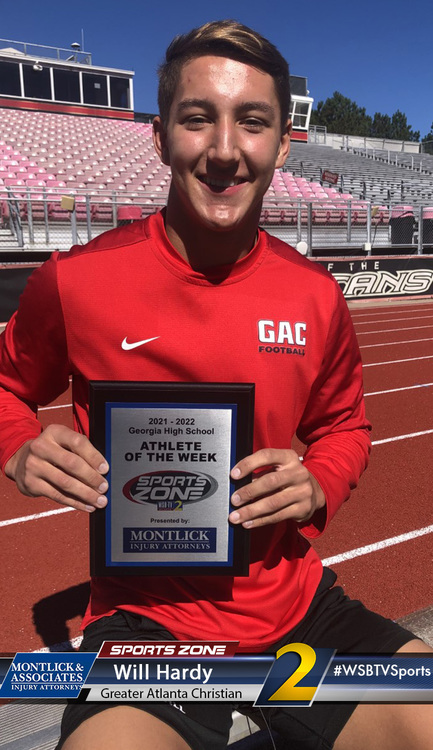 Talented high school athlete holding an "Athlete of the Week" award from a local news station, wearing a red athletic jersey and smiling at the camera while sitting on a red running track on a sunny day.