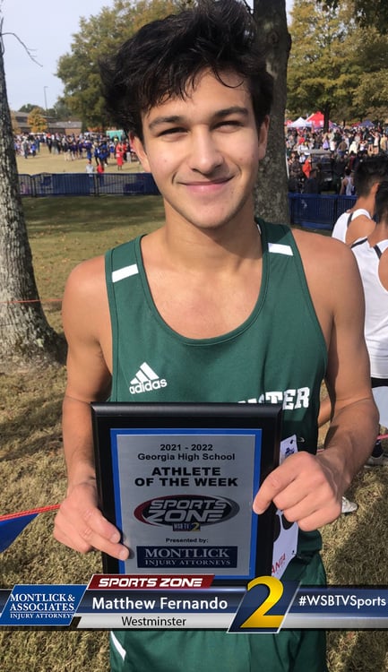Young male athlete smiles holding Athlete of the Week award at high school cross country meet.