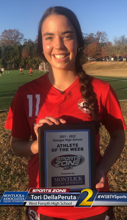 Young female athlete of the week smiling and holding award plaque on sports field, wearing red uniform.