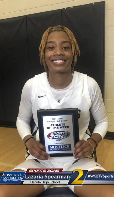 Young smiling athlete holds "Athlete of the Week" award from Sports Zone magazine while sitting at desk.